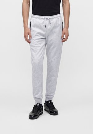 Homme portant un pantalon de jogging gris clair avec taille à cordon et poches latérales zippées noires, associé à des baskets noires.