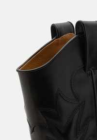 Černá kožená kotníková bota s hladkým povrchem, zdobená dekorativním prošíváním a hnědým interiérem. Dvojité poutka pro snadné obouvání.