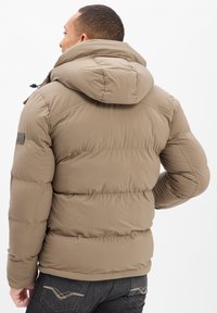 Cazadora acolchada beige con capucha, diseño acolchado y puños elásticos. Tejido suave con una textura lisa y un sutil logo en la manga.