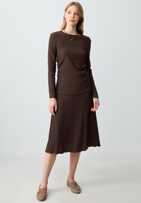 Bruine geribbelde lange mouwen top in combinatie met een bruine midi rok. Het materiaal lijkt textuur te hebben, met een golvende zoom en zachte, neutraal gekleurde loafers.