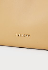 Tan textured leather tote featuring embossed "TED BAKER" logo in gold. Smooth finish with a rectangular shape and minimalistic design.