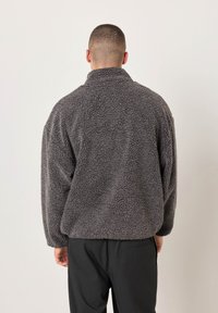 Žmogus matomas iš nugaros, dėvintis tamsiai pilką tekstūruotą vilnos striukę ir juodas kelnes, stovintis prieš paprastą šviesų foną.