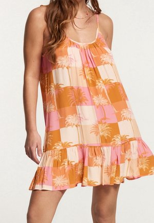Mujer con vestido sin mangas a cuadros rosas y naranjas con estampados de palmeras y bajo con volantes frente a un fondo liso y claro.