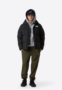 Fekete steppelt dzseki kapucnival, alatta szürke kapucnis pulóver, olívazöld nadrág és fekete bakancsok. A dzsekin és a sapkán logók láthatók.