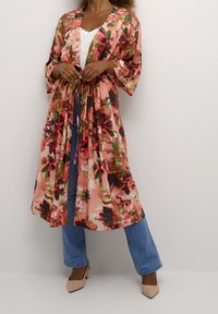 Lång, blommig kimono i rosa och gröna nyanser med knytning i midjan, vida ärmar och en genomskinlig textur, i kombination med ljus denim och beige skor.