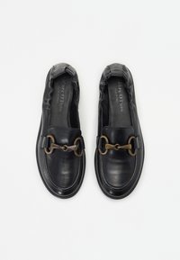 Zwarte leren instapschoenen met gegatherde randen, voorzien van een goudkleurige kettingaccent en een gladde textuur. Platte zool, casual ontwerp.