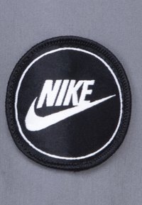 Zwarte cirkelvormige patch met witte "Nike" tekst en swooshlogo op een grijze textielachtergrond. De patch heeft een gestikt rand.