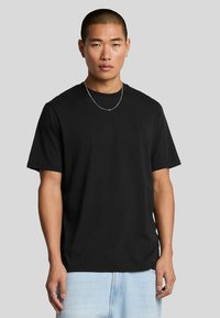 T-shirt nera in cotone con maniche corte, collo a giro e un discreto logo ricamato sul petto. Il modello indossa jeans azzurri chiari e gioielli argentati.