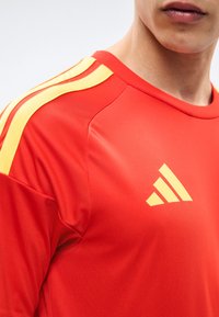 T-shirt de sport rouge avec des bandes jaunes sur l'épaule et un logo Adidas jaune sur la poitrine, porté par une personne visible du cou jusqu'au milieu du torse.