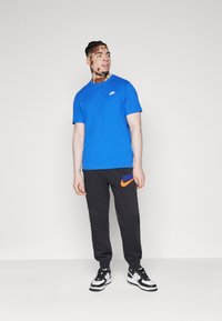 T-shirt blu a maniche corte con logo Nike bianco, abbinato a pantaloni della tuta neri con logo Nike arancione. Sneakers in bianco e nero completano il look.