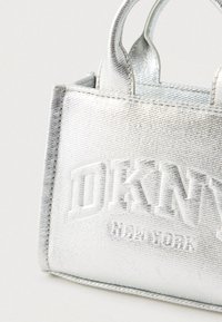 Hõbedase metallikvärviga DKNY kandekott reljeefse logoga ja lühikeste käepidemetega, taustal hele ühtlane pind.