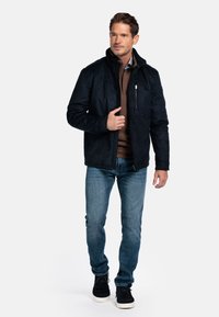 Man in een zwart ritsjack, bruine pullover, blauwe spijkerbroek en zwarte casual schoenen, staand voor een witte achtergrond.