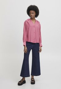 Camicia blouse rosa con maniche lunghe, caratterizzata da una scollatura a V e spalle arricciate. Abbinata a pantaloni in denim scuro a gamba ampia. Sandali platform neri.