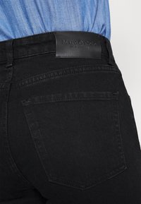 Pantalones vaqueros de denim negro con una textura suave, que presentan una etiqueta de cuero con "Marc O'Polo" en la cintura y bolsillos traseros estándar.
