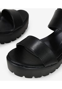 Las sandalias de plataforma negras tienen una textura de cuero suave, dos correas anchas que cruzan el pie y una suela gruesa y estriada para mayor estabilidad.