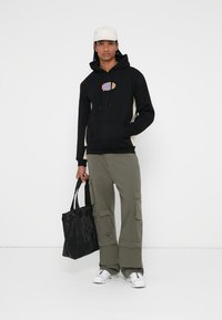Sudadera con capucha negra y paneles laterales beigs, con un logo multicolor. Pantalones cargo oliva y una bolsa de mano negra completan el atuendo.