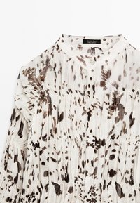 Blouse boutonnée avec un col en V, réalisée dans un tissu léger texturé de couleur crème avec des motifs éclaboussés marron et des volants à étages.