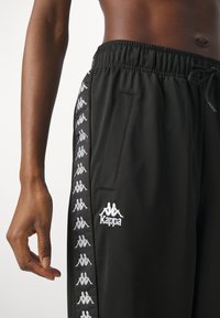 Pantaloni atletici neri con cintura elastica, tasca laterale e logo Kappa. Presenta un motivo con logo bianco ripetuto lungo il lato.