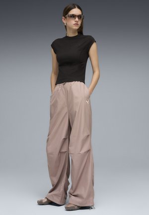 Jeune femme portant un haut noir sans manches, un pantalon beige ample avec des poches, des lunettes de soleil et des chaussures texturées, debout les mains dans les poches.