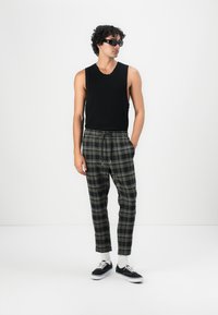 Schwarzes ärmelloses Oberteil, karierte Hose in Grüntönen und Grau, mit Kordelzugbund und schwarzen Turnschuhen. Zubehör umfasst dunkle Sonnenbrille.