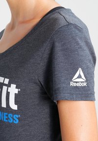 Šedé sportovní tričko s výstřihem do V, krátkými rukávy a výrazným bílým logem Reebok na pravém rameni.