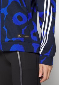 Sudadera azul marino con grandes patrones florales azules, acentos en blanco de tres rayas en las mangas y un logo dorado de Adidas marimekko en el lateral.