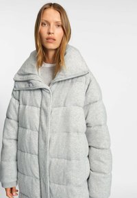 Abrigo acolchado gris claro con un cuello alto, que presenta un cierre con cremallera, botones a presión y textura acolchada. Tejido suave con un patrón sutil.