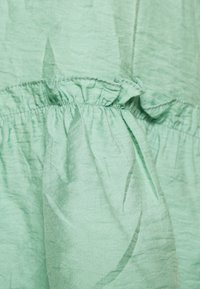 Světlý zelený, pomačkaný materiál s volánkovým švem a nabranou strukturou. Měkký materiál s lehkým vzhledem. Žádné viditelné vzory ani kovové prvky.