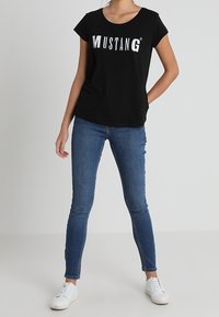 Kobieta ubrana w czarną koszulkę z logo Mustang, niebieskie obcisłe dżinsy i białe sneakersy, stojąca z jedną ręką na biodrze.