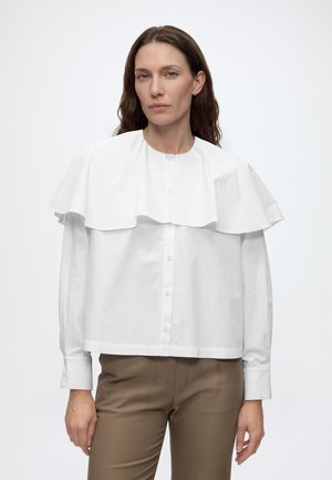 Woman with long brown hair wearing a white buttoned shirt with a large cape collar and brown pants standing against a plain background.
