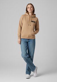 Felpa beige con testo nero "AMOUR", tasca a marsupio, cordini, abbinata a jeans azzurri chiari e sneakers bianche. Texture liscia e morbida.