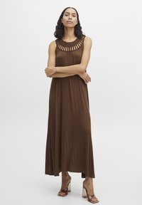 Robe maxi sans manches marron avec un détail tissé au niveau du col. Tissu doux, silhouette fluide et ourlet droit. Chaussures à talons hauts transparentes.