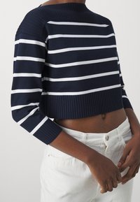 Maglione cropped a righe blu navy e bianche con scollatura e polsini a coste, abbinato a pantaloni bianchi. Il tessuto sembra morbido e elasticizzato.
