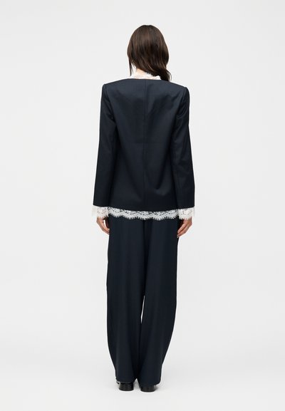 Γυναίκα που στέκεται με την πλάτη, φορά μαύρο σακάκι και φαρδιά παντελόνια με λευκή δαντέλα ορατή στα μανίκια και το τελείωμα.