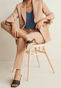 Beige přizpůsobený blejzr a kalhoty, vrstvené přes modré tričko. Texturované černé sandály na podpatku. Dřevěná židle, neutrální pozadí.