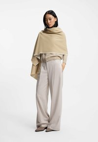 En beige sjal hænger over en lys sweater, parret med lysegrå, vidde bukser. Sjalet har en kontrastkantdetalje.