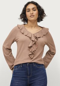 Langarmshirt van lichtbruin textiel, met een gerimpelde halslijn en voorlaatste detail, gecombineerd met donkerblauwe denim jeans.