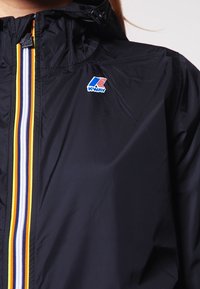 Czarna, lekka kurtka z kapturem, z centralnym paskowanym wzorem w kolorach żółtym, białym i niebieskim. Zawiera logo K-WAY.