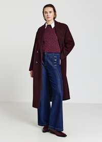 Cappotto in lana bordeaux con grandi bottoni, sovrapposto a un maglione a trecce. Abbinato a jeans in denim a vita alta e a gamba svasata e stivaletti bordeaux.