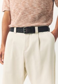 Cintura nera in pelle con fibbia di metallo lucido, indossata sopra pantaloni plissettati color crema e un top marrone lavorato a maglia con trama.