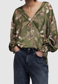 Blouse à manches longues de couleur olive avec des motifs floraux et d'oiseaux. Caractérisée par un col en V profond et des poignets froncés. Associée à un jean bleu foncé.