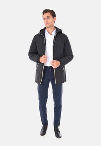 Uomo che indossa una giacca nera con cappuccio sopra una camicia bianca, pantaloni blu marino e scarpe nere, in piedi contro uno sfondo chiaro e uniforme.