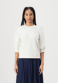 Cream sweatshirt with puffed sleeves, round neckline, and textured fabric, paired with a dark pleated skirt. Minimalist, contemporary design.