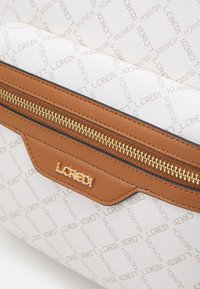 Sac à main blanc avec une finition texturée, présentant un accent de zip marron et un logo doré. Le design comprend un motif de texte de marque répétitif.