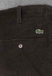 Pantalon en velours côtelé marron avec des côtes verticales, arborant un petit logo Lacoste vert et un bouton en métal gravé "Lacoste".