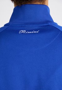 Mėlyna polo marškinėliai su aukštu apykakle. Jie turi lygų paviršių ir siuvinėtą baltą "Rimini" logotipą ant galinės apykaklės.