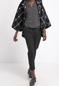 Person, die einen schwarzen karierten Umhang mit Reißverschluss, eine gemusterte Bluse, schwarze Cropped-Hosen und schwarze Plateau-Loafer mit Reißverschlussdetails trägt.