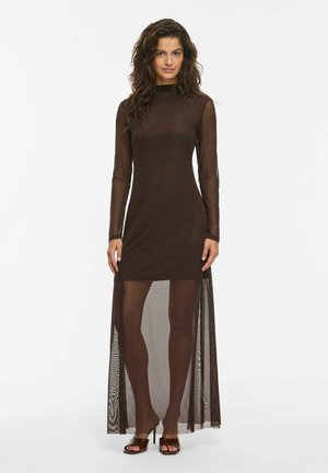 Une femme présente une robe ajustée brun foncé, à manches longues transparentes et recouverte d’un voile en résille jusqu’au sol, sur un fond blanc uni.