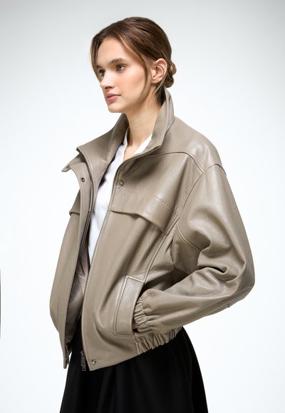 Junge Frau trägt eine braune Lederjacke mit hohem Kragen und elastischen Bündchen, steht im Profil mit den Händen in den Taschen vor einem weißen Hintergrund.