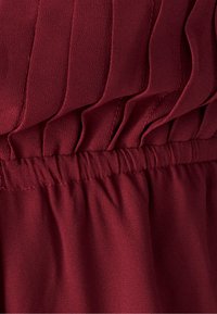 Temno rdeča bluza z navpičnimi naborki, z elastiko v pasu, gladke teksture in lahkega materiala.